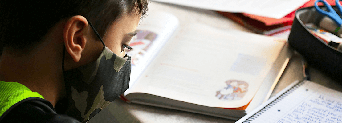 A young boy wearing a facemask writes in a school book