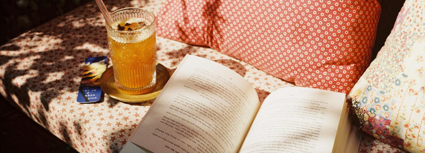 Picture of an open book on a bed or couch with a glass of iced tea next to the book and a pillow in the background.
