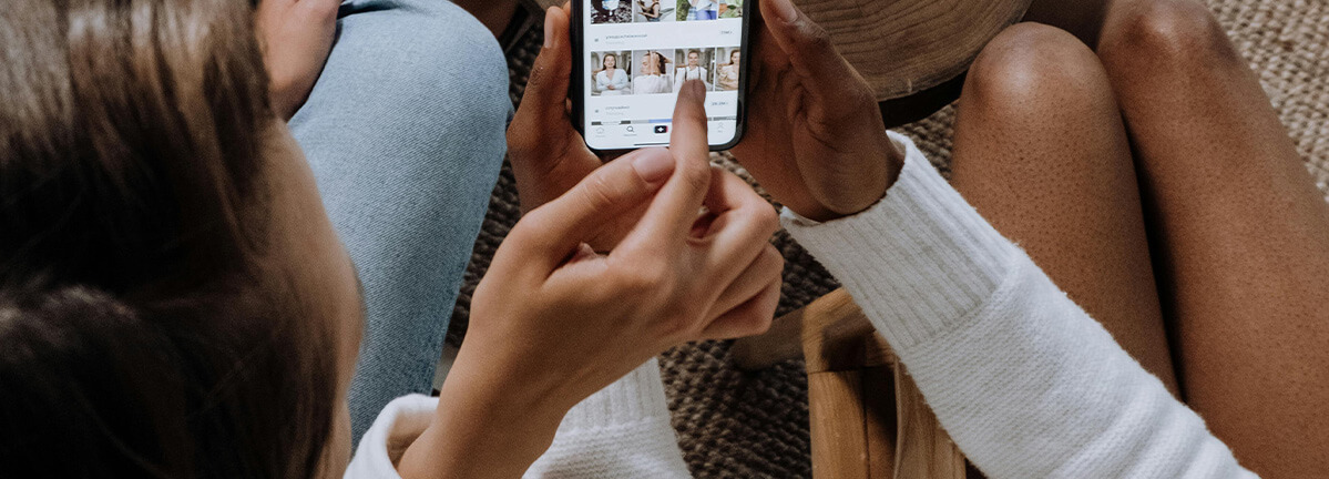 Two people look at a phone together while they sit on a couch. One person holds the phone in their hand while the other uses their finger to scroll.